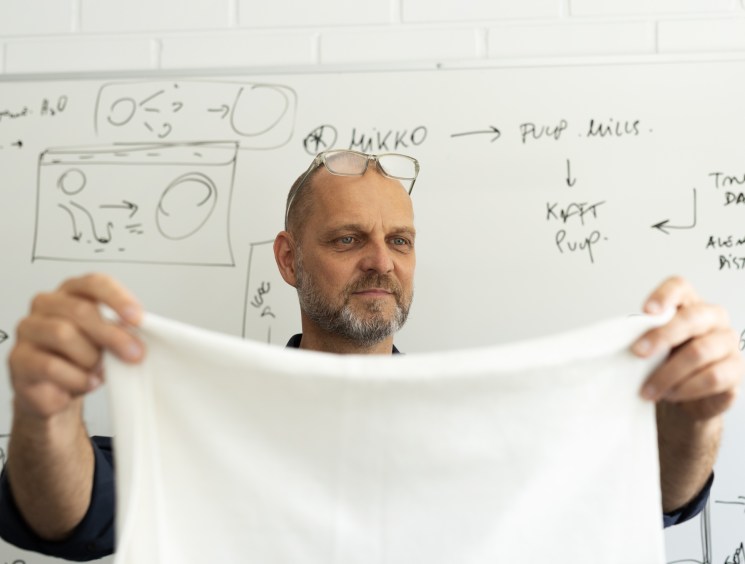 Man with glasses and beard holding white material sample in front of whiteboard with diagrams