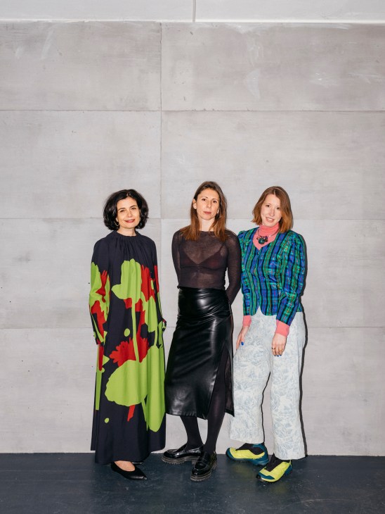 Three women standing together in contemporary fashion against a concrete wall