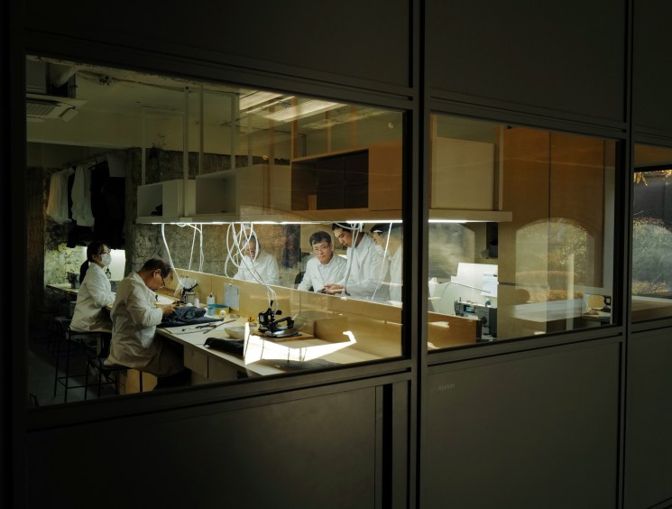Four people in white lab coats working at tables with equipment in a well-lit workshop space viewed through glass