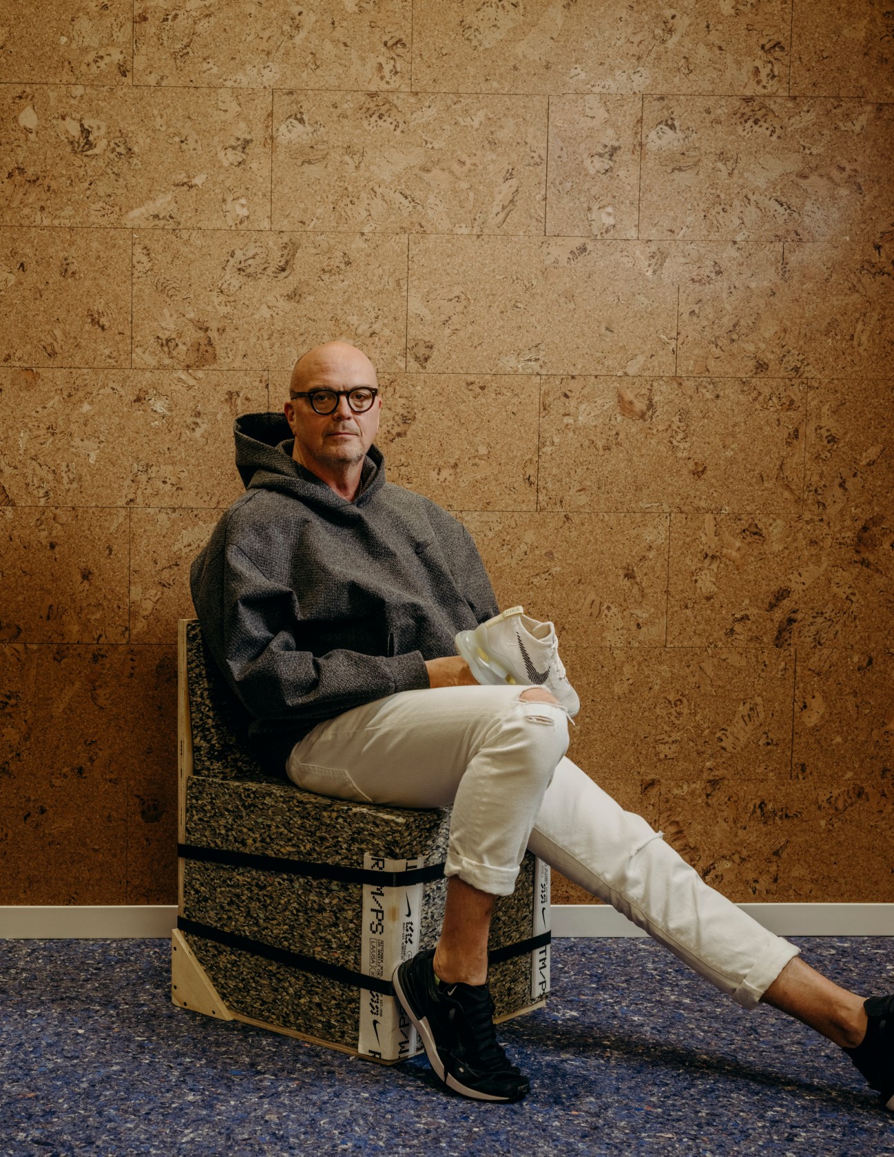 John Hoke III sitting casually on stacked boxes wearing glasses, gray hoodie and white pants