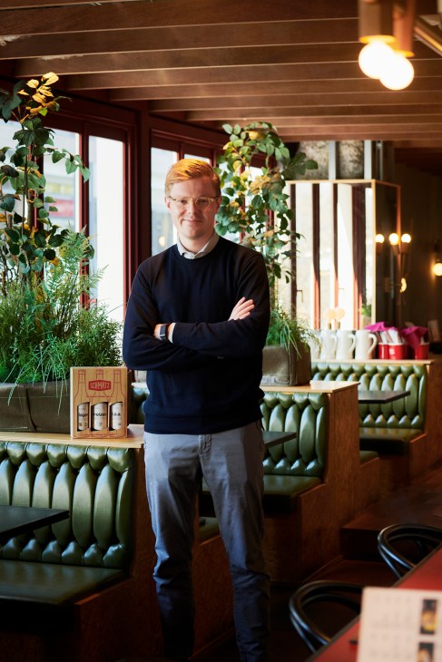 Marc Luetten standing with crossed arms in Schmatz restaurant interior with green booth seating and plants