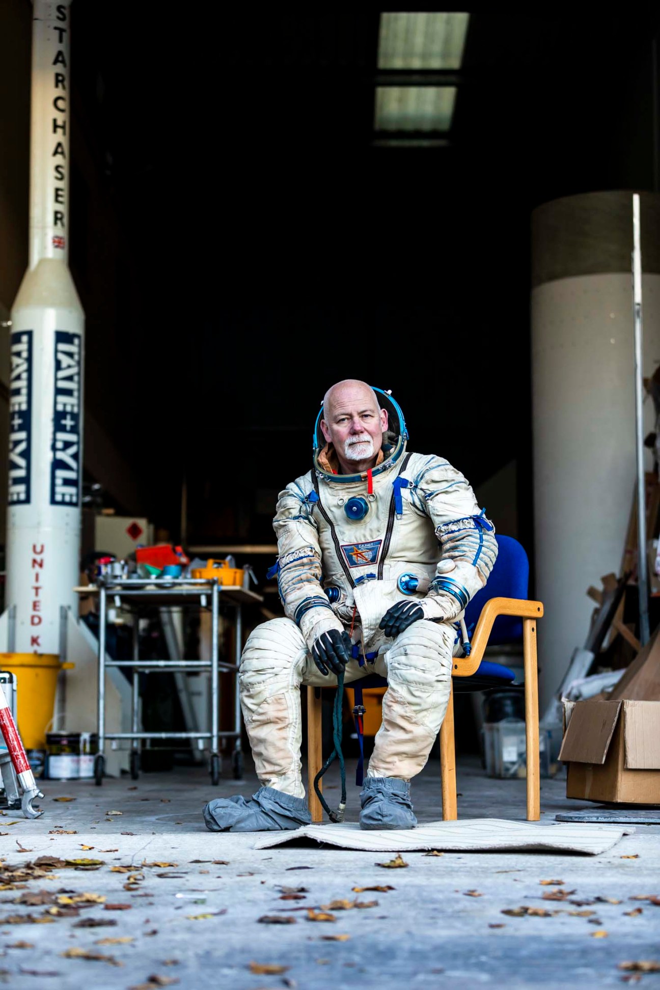 Man in white spacesuit with blue trim sitting on wooden chair next to rocket and scaffolding in industrial setting.