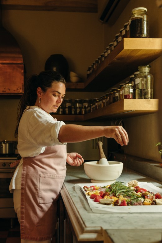 Woman in white shirt and pink apron using mortar and pestle in rustic kitchen with jars on wooden shelves