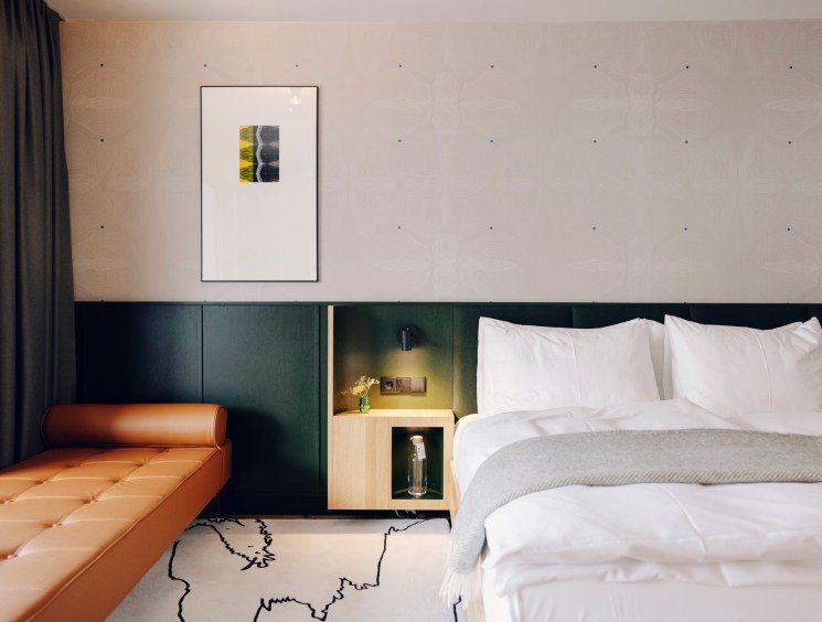 Hotel room with white bedding, orange leather bench, dark green headboard wall, and framed artwork