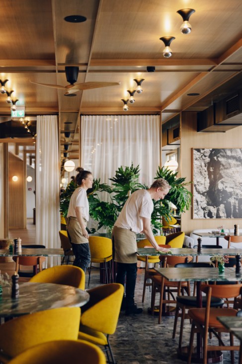 Two staff members in beige aprons setting up a restaurant dining room with yellow chairs and large plants