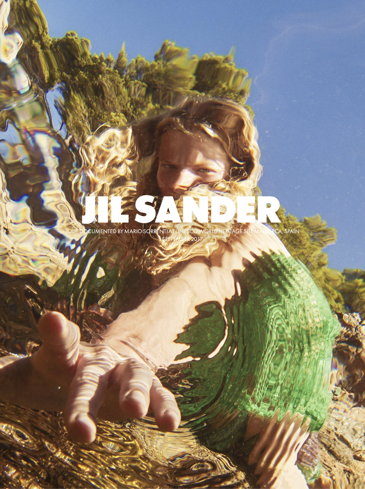 Model in white clothing among golden sculptural elements and green foliage under blue sky for Jil Sander campaign