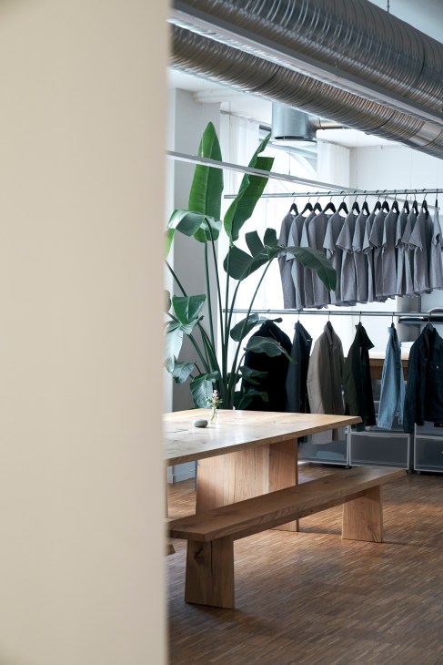 Modern clothing store interior with wooden bench, exposed ductwork, plants, and racks of gray garments