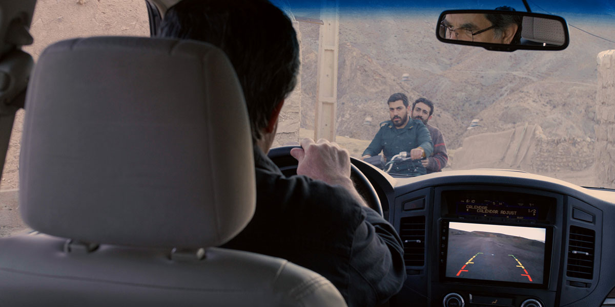 View from inside a car showing two men on a motorcycle outside in an arid landscape