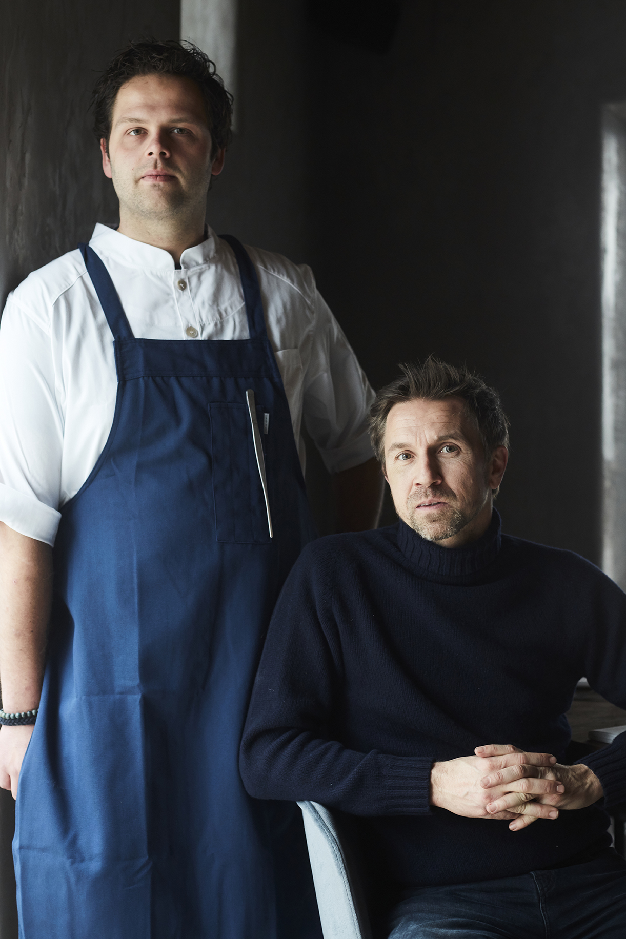 Two men posing for portrait, one standing wearing chef's apron and white shirt, one seated wearing black turtleneck