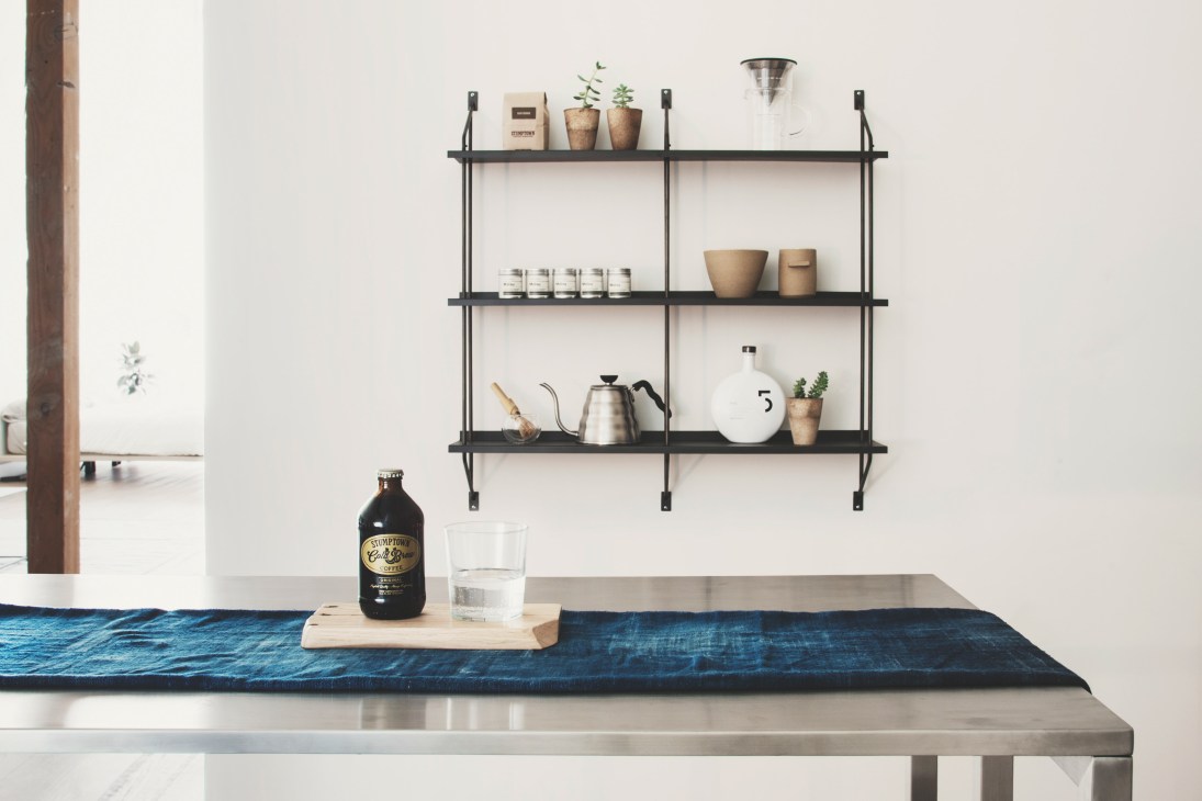 Black metal shelving unit displaying potted plants, coffee equipment, candles and home goods on a white wall above a counter with Stumptown Coffee bottle.
