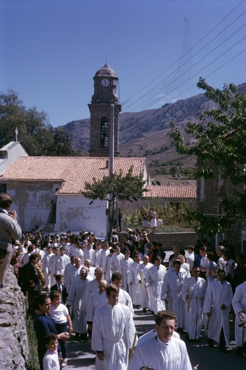 Large religious procession of people in white robes walking through Mediterranean village with bell tower
