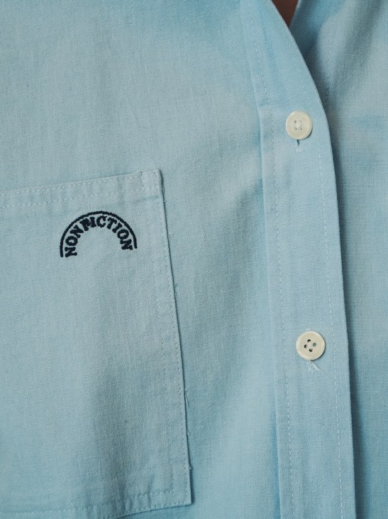 Close-up of pale blue chambray shirt with subtle Nonfiction brand logo embroidered on chest pocket