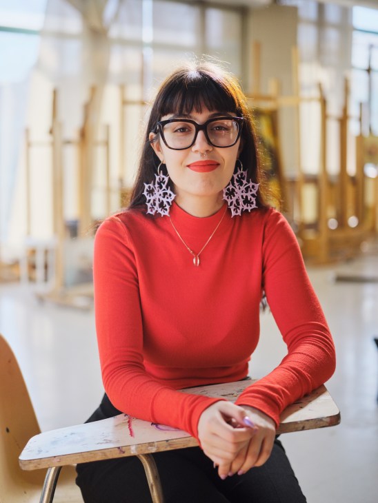 Young woman with glasses and bangs wearing red sweater and decorative earrings sitting at wooden table
