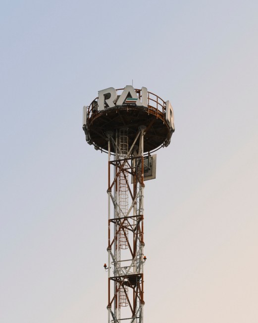 Tall metal lattice transmission tower with RAI lettering on circular platform against pale sky