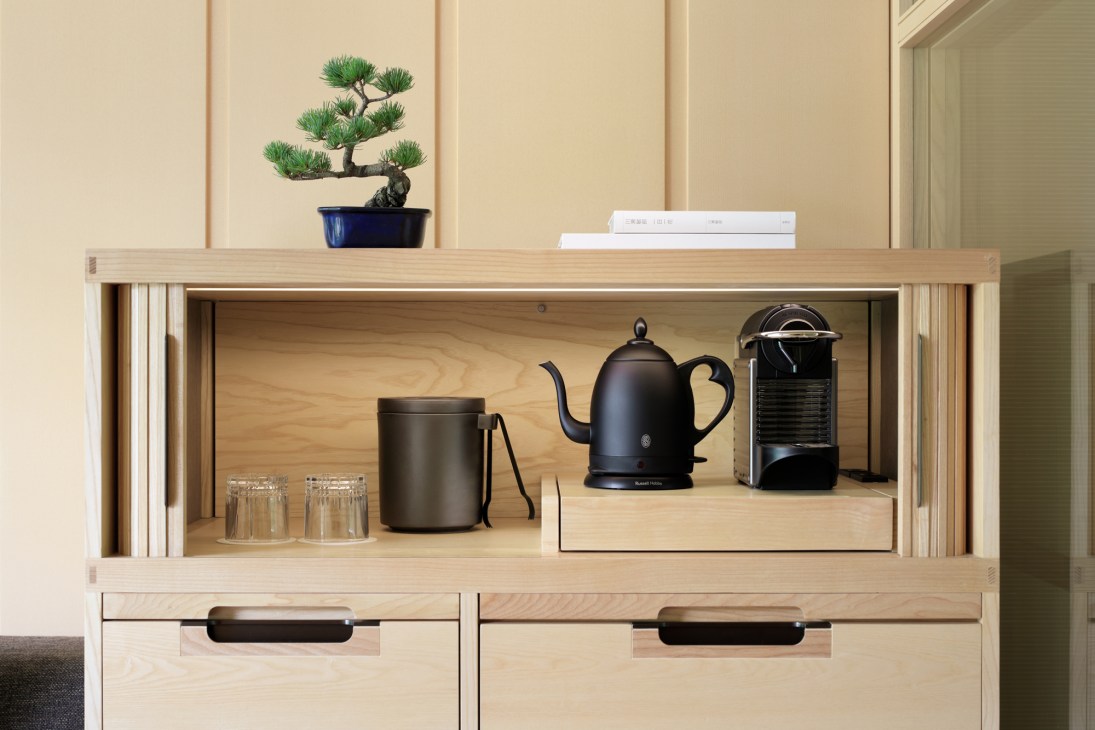 Light wood shelving unit displaying a bonsai tree, black tea kettle, coffee machine, and stacked books.