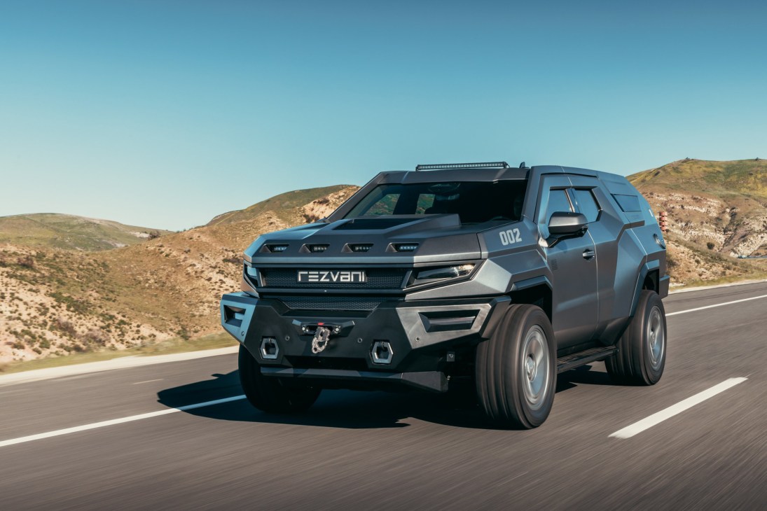 Gray armored Rezvani Vengeance SUV driving on desert highway with rugged hills in background