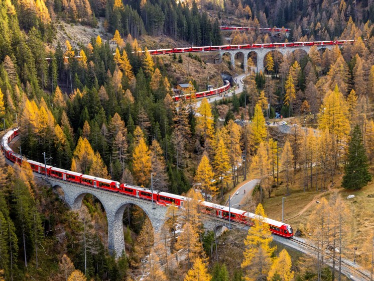 Red Rhätische Bahn train crossing stone viaduct through autumn forest in Swiss mountains