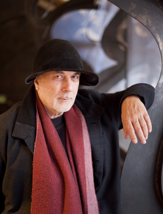 Ron Arad wearing black hat and red scarf, standing next to curved sculptural furniture piece in his design studio