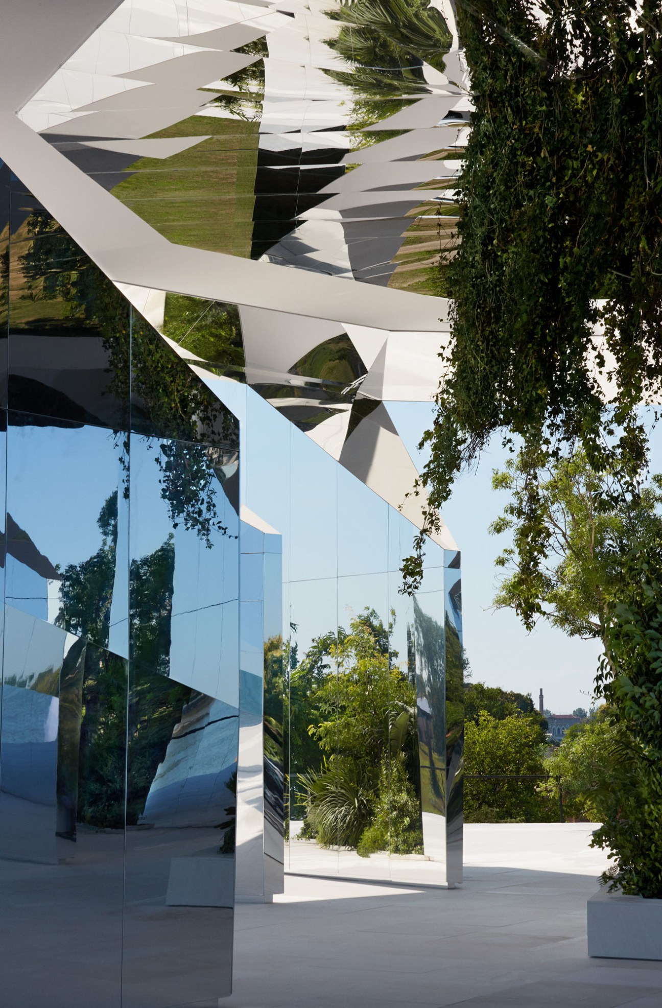 Angular mirrored pavilion installation by Doug Aitken on Venetian island with geometric white panels and greenery