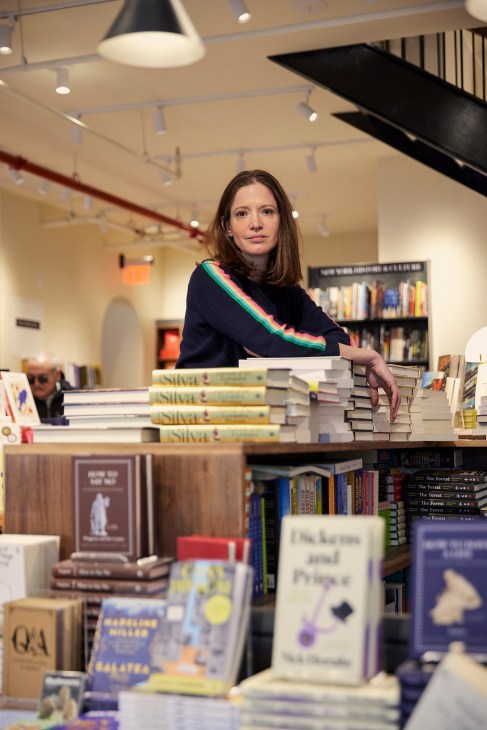 Woman with brown hair wearing dark sweater with rainbow stripe standing among stacks of books in bookstore