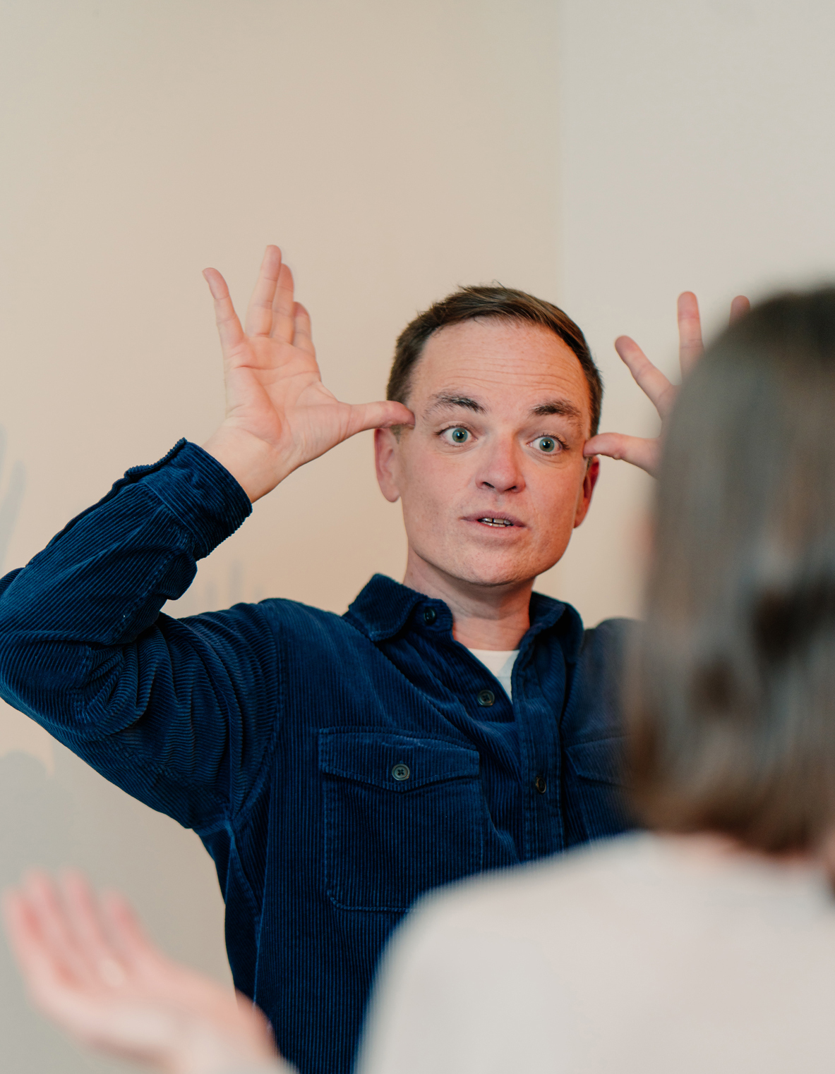 Man in blue corduroy shirt making exaggerated facial expressions with hands raised during improv class