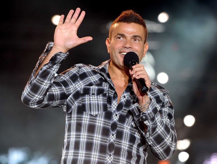 Amr Diab performing on stage with microphone, wearing plaid shirt and waving to audience