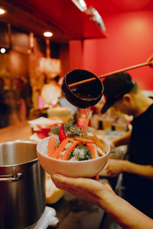Hand holding a bowl of colorful Asian noodle dish with vegetables and dark sauce being poured from ladle