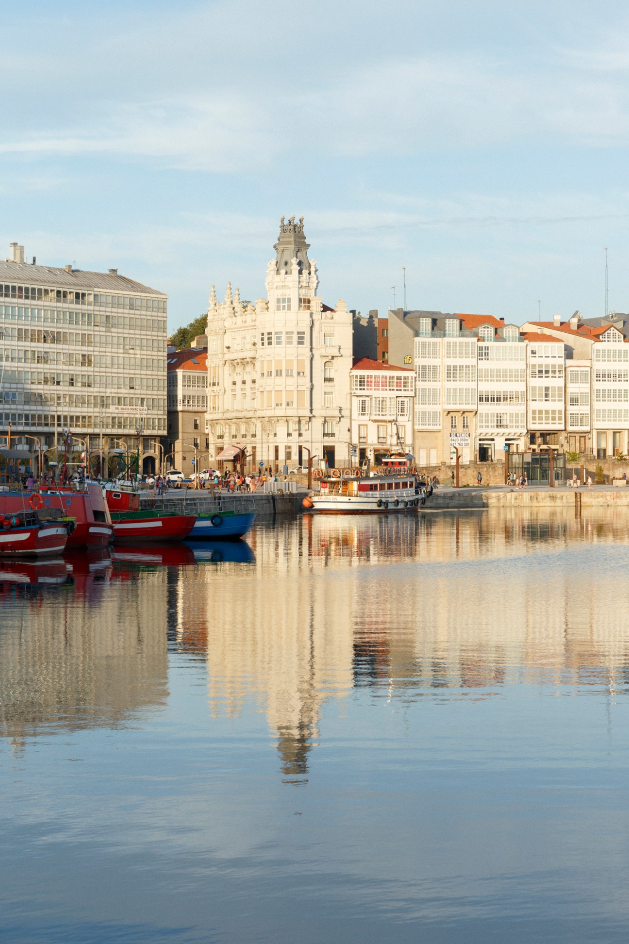 A Coruña's waterfront with colorful fishing boats, historic architecture, and a prominent cream-colored building reflected in calm water.