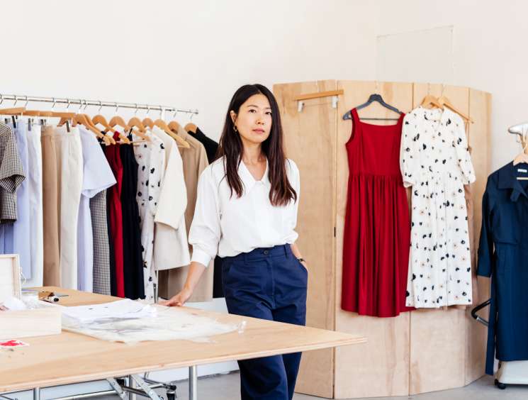 Jeana Sohn stands in her studio with clothing designs on hangers and work materials on a table.