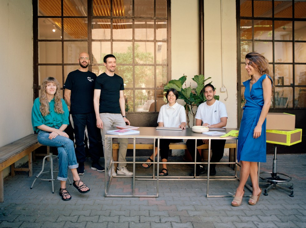 Studio Vedèt team of six people posing in a bright workspace with large industrial windows and plants.