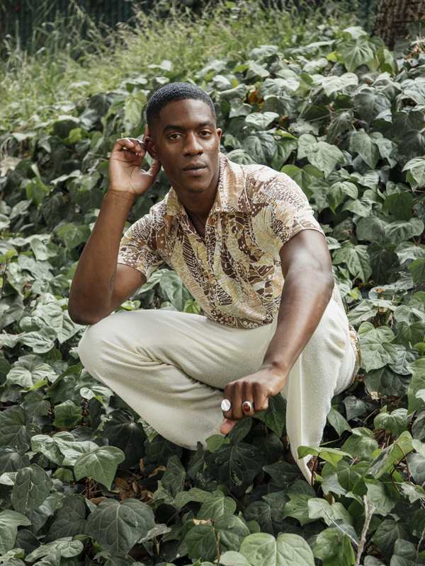 Man wearing patterned shirt sitting cross-legged among green leafy plants