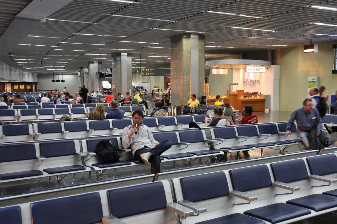 Airport terminal waiting area with blue seating and passengers, featuring modern ceiling design