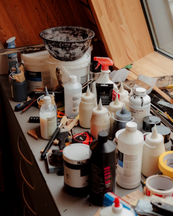 Artist's studio workspace with paint bottles, brushes, and art supplies scattered on a white table