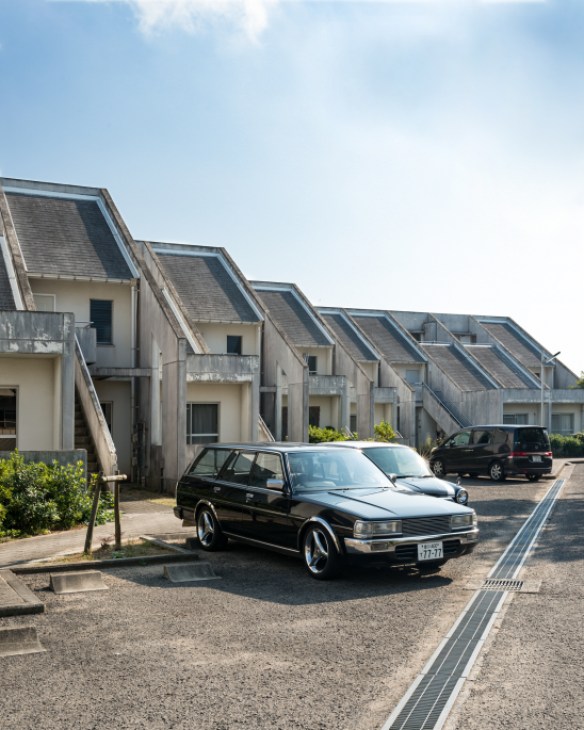 A black Nissan Bluebird estate car parked in front of modern residential houses with distinctive angled roofs
