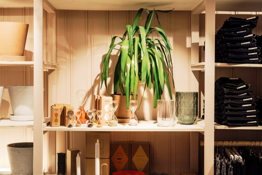 Wooden shelving displaying Hay products including hourglasses, glassware, and a potted trailing plant in a minimalist retail space.