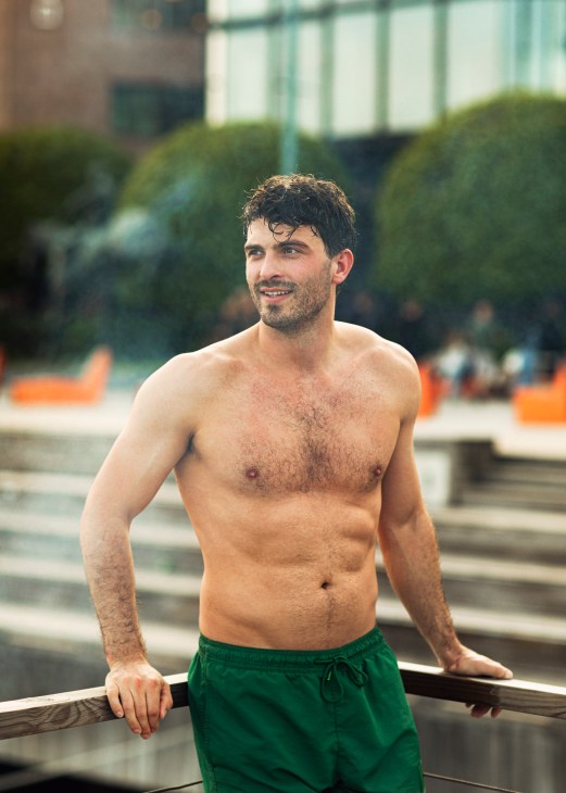 Shirtless man in green swim shorts leaning against railing near outdoor pool or waterfront area