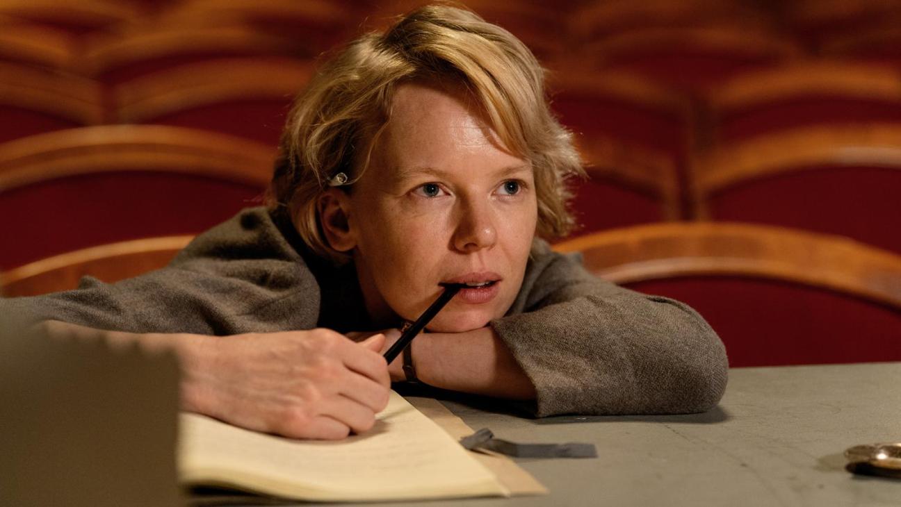 A young person with reddish-blonde hair sits at a desk, thoughtfully holding a pen to their mouth while looking at papers.