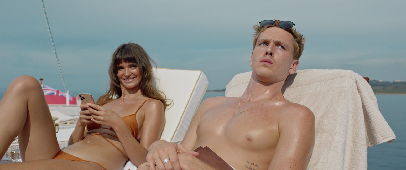 Two people lounging on deck chairs aboard a luxury yacht, woman in bikini using phone, shirtless man beside her