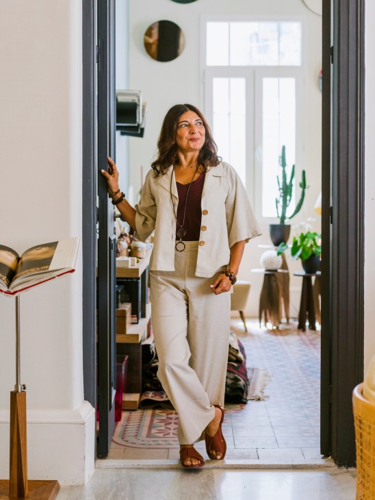Nada Debs stands in her studio doorway wearing a cream linen suit, surrounded by plants and modern furnishings.