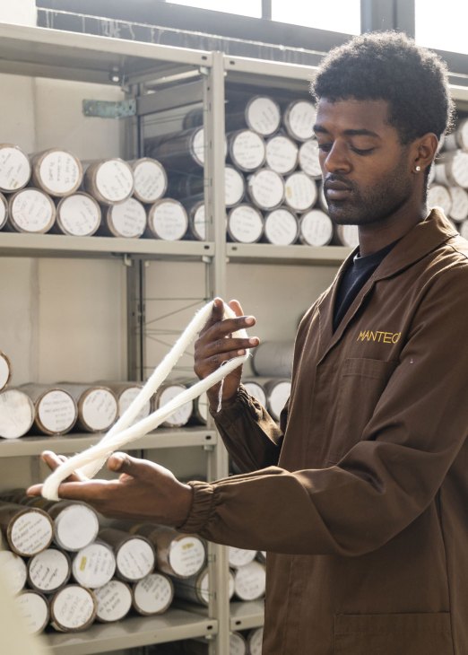 Worker at Manteco factory examining white yarn with shelves of fabric rolls in background