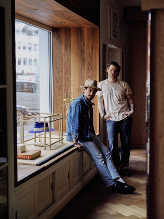 Two men pose in a modern wooden interior, one sitting on a window ledge wearing a hat, the other standing beside him