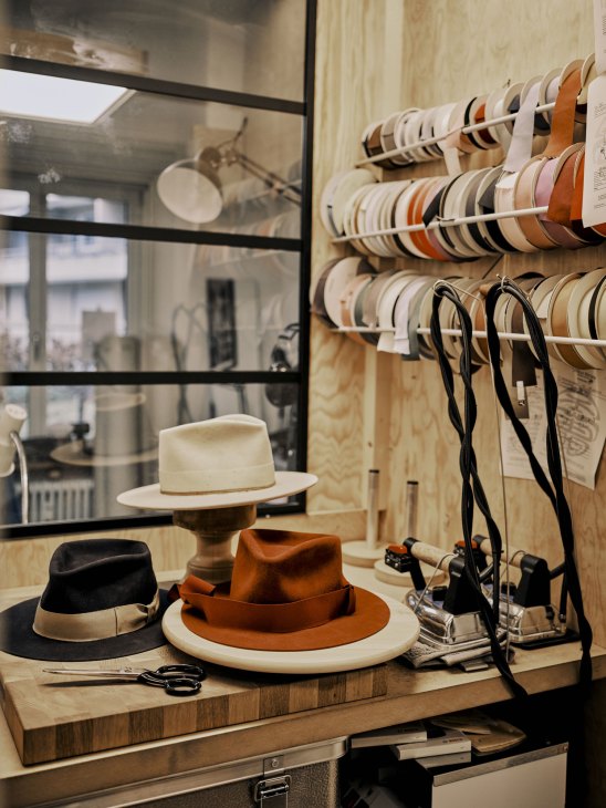 Hat-making workshop with finished fedoras on wooden workbench, colorful hat bands on wall rack, and crafting tools