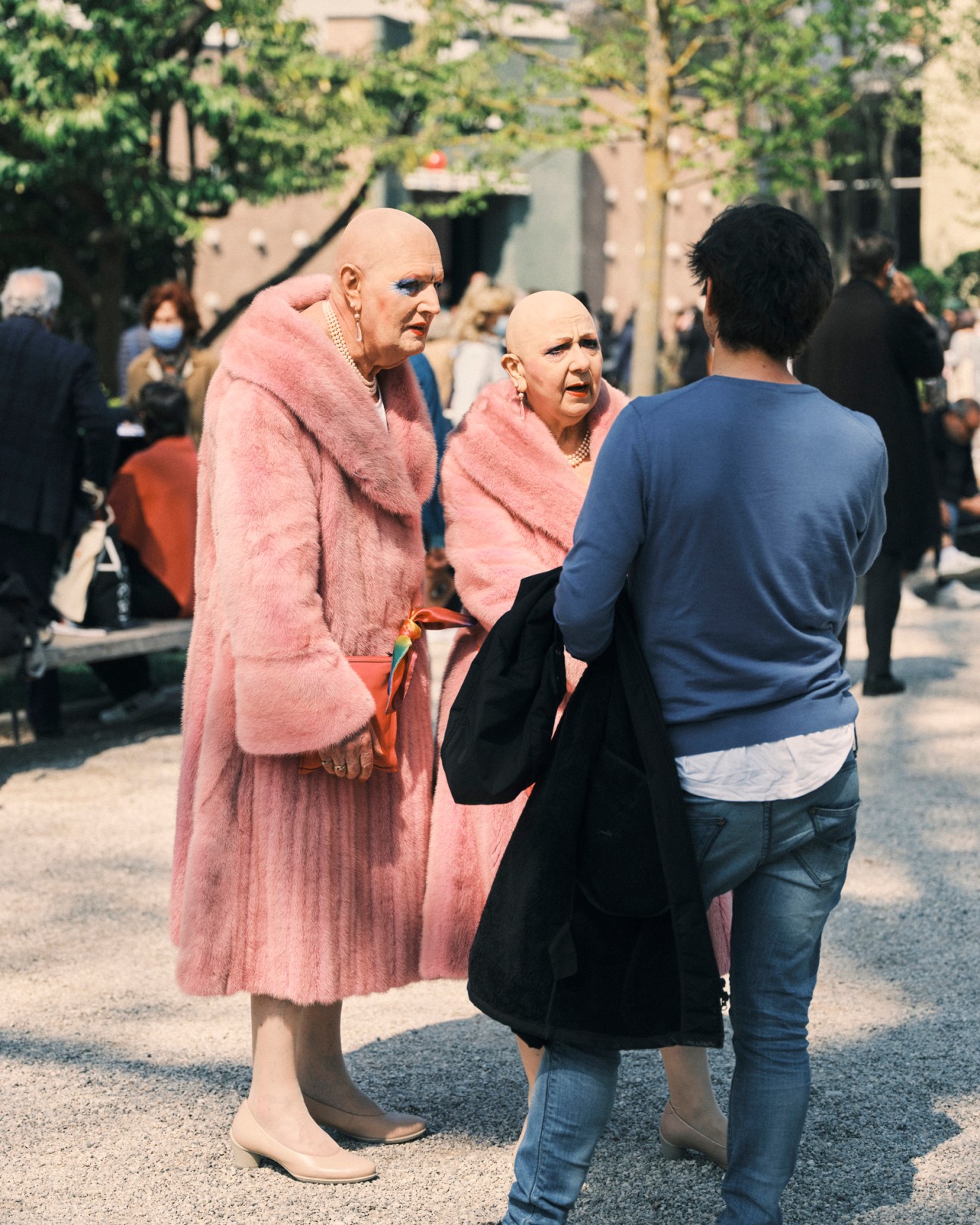 Two bald figures wearing matching pink outfits converse with a person in a blue shirt in a sunlit courtyard with crowds and historic buildings in the background.