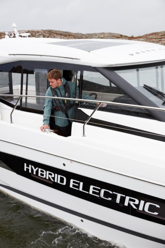 Man in green jacket operating white hybrid electric boat with 'HYBRID ELECTRIC' text on hull at Swedish test facility