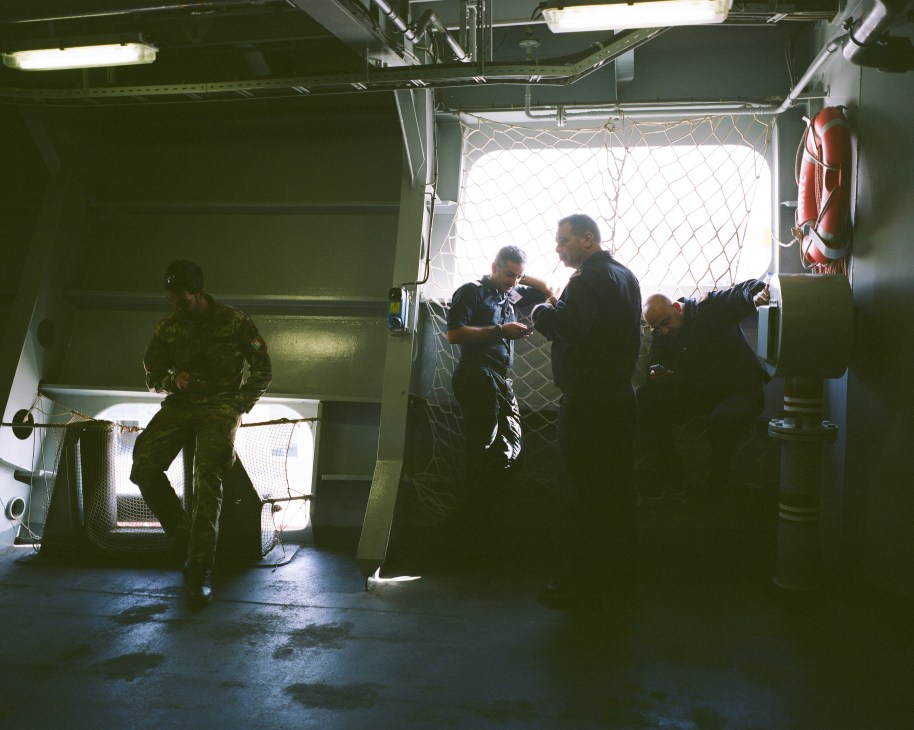 Italian sailors in military uniforms stand in a corridor of the Cavour aircraft carrier near a ship seal emblem.