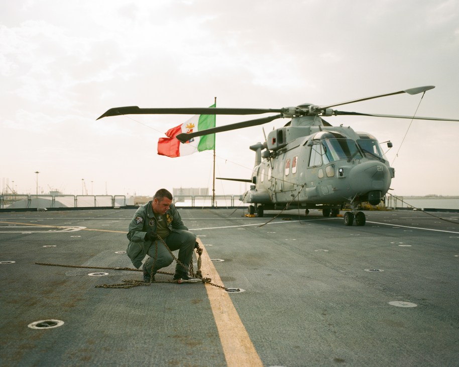 A flight deck crewman crouches on the Cavour's deck removing a cable, with an Italian military helicopter and flag visible in the background.