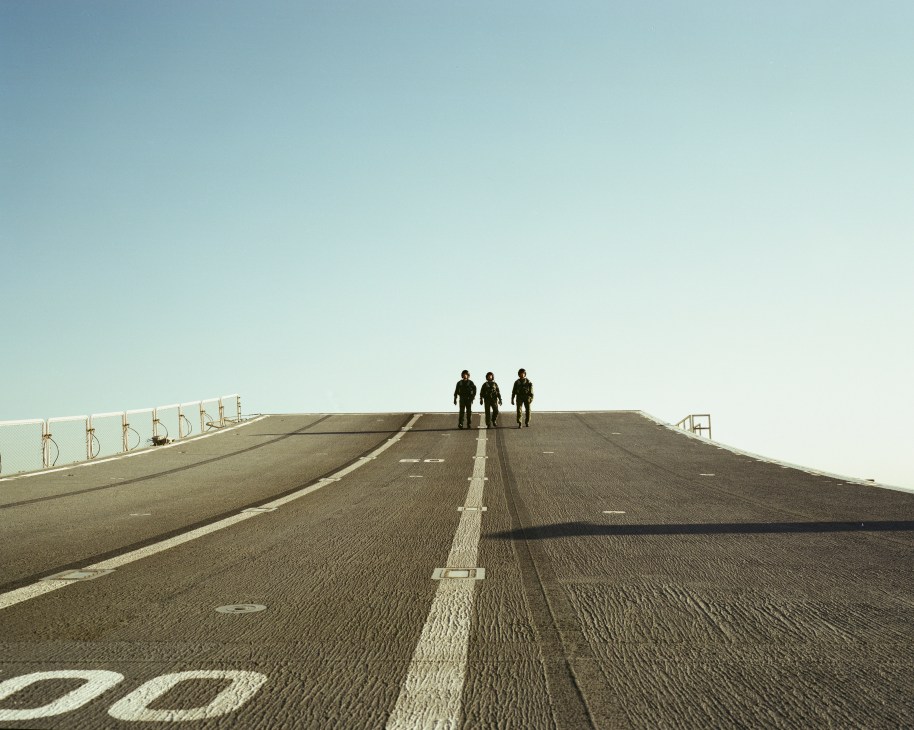 Three military personnel stand on the angled takeoff ramp of an aircraft carrier flight deck.