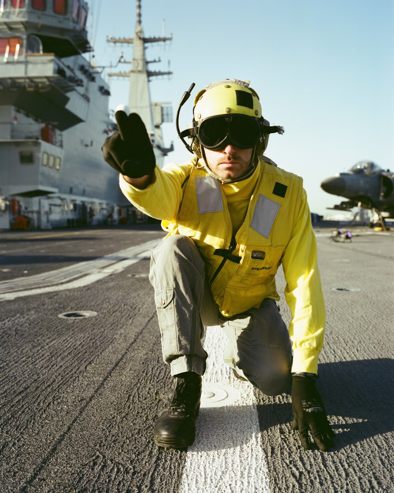 Flight deck crew member in yellow vest and helmet giving hand signal on aircraft carrier flight deck.