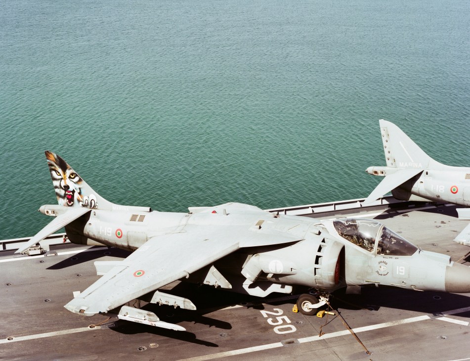 AV-8B Harrier II fighter jets parked on the Cavour aircraft carrier flight deck with turquoise sea beyond.