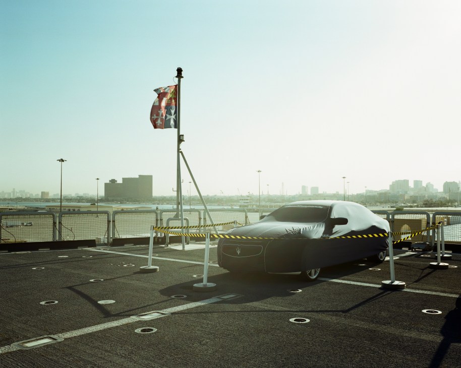 A covered Maserati luxury car sits on the Cavour aircraft carrier's flight deck with an Italian flag and cityscape visible in the background.